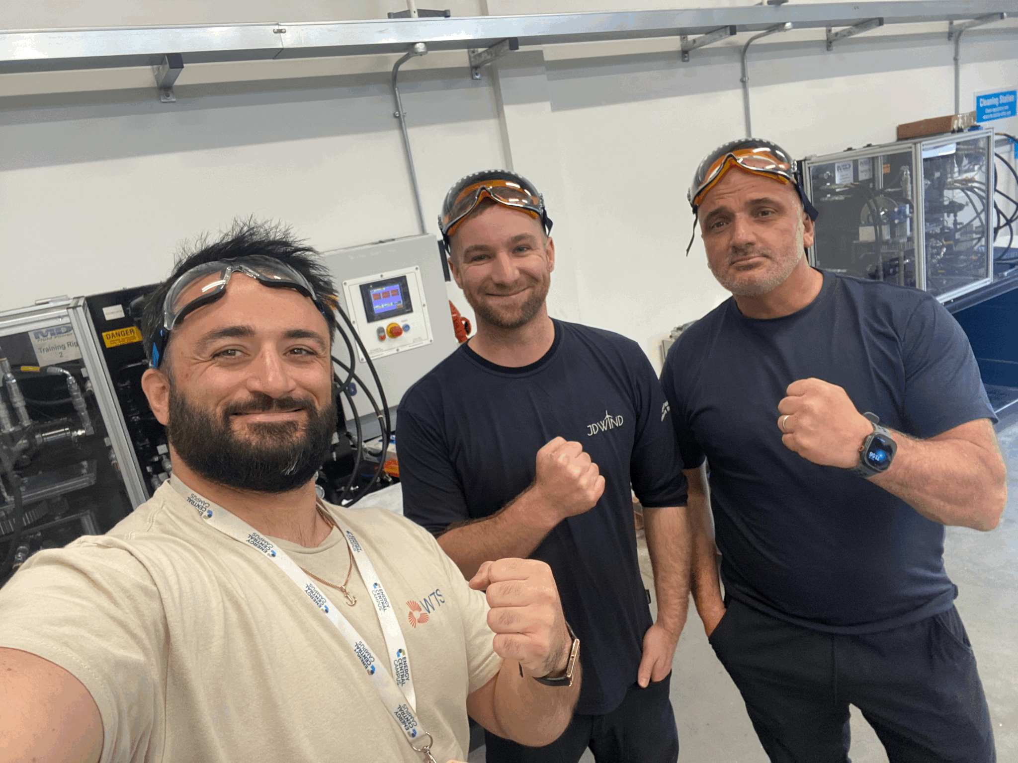 Three wind turbine technicians in safety goggles, fists raised and smiling, in the WTS training workshop with hydraulic and electrical rigs behind them. One wears a JD Wind T-shirt, one a WTS T-shirt.