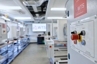 A spacious electrical workshop featuring several control panels, training rigs, and a TV displaying the WTS brand.
