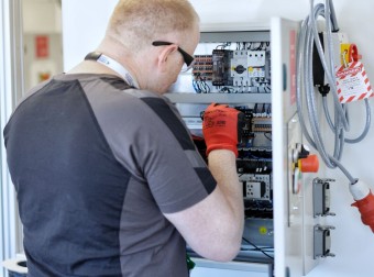 A man in a black shirt and red gloves is focused on working on a control panel.