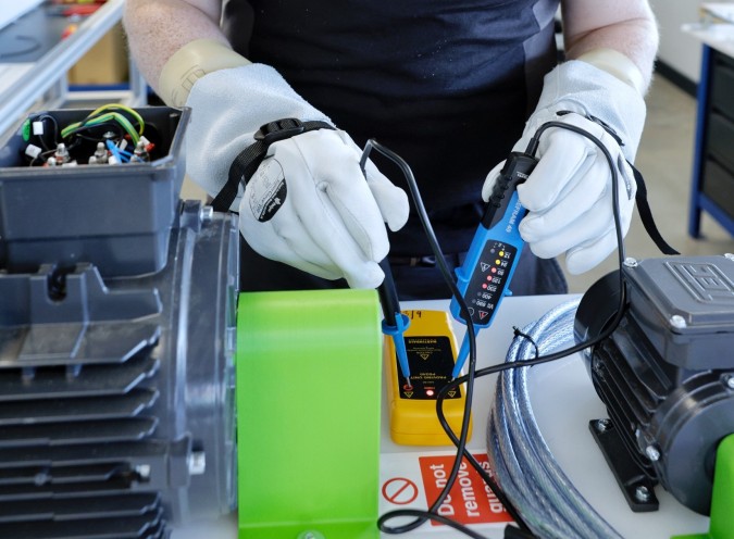 A student wearing gloves, using electrical testing equipment with a tool in hand.