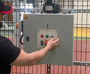 A training instructor in a black shirt and red hat stands beside a metal box, showcasing electrical equipment.