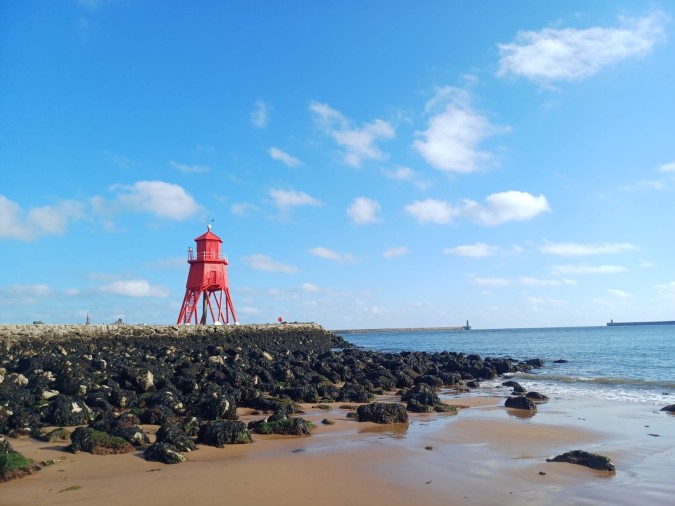 A red lighthouse, The Groyne, stands on the beach beside rocky formations in South Shields, UK.