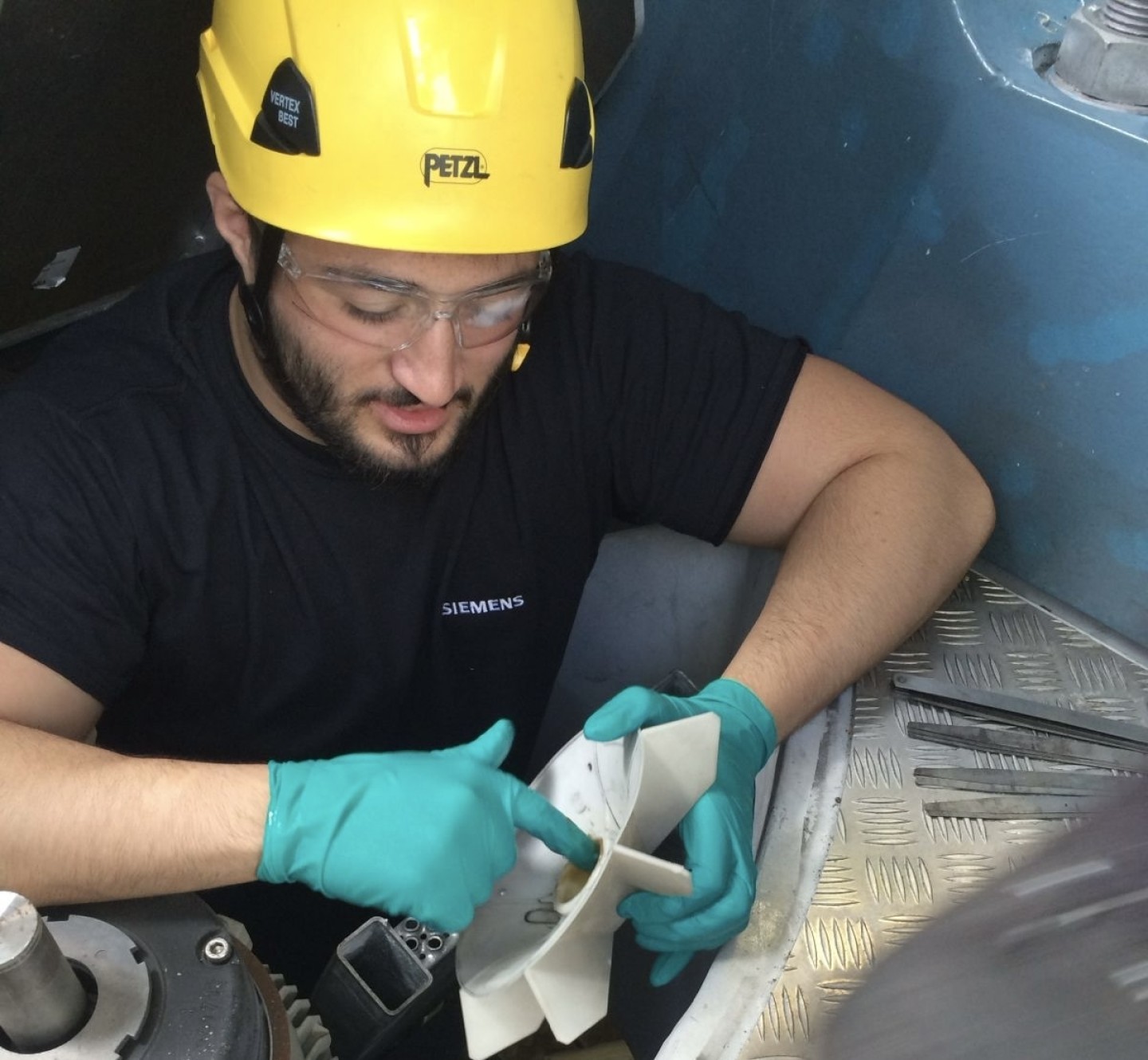 A person in safety gear, including a yellow helmet and gloves, checks machinery inside an industrial setting. Tools lie on a metal grid floor nearby.