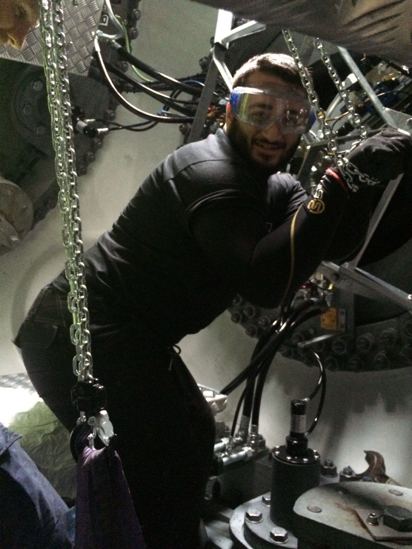 A person wearing safety goggles and gloves works in a mechanical environment, gripping chains and smiling. The area is filled with machinery and metal parts.